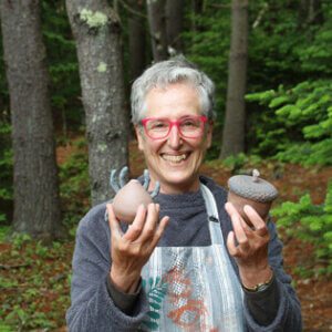 A smiling artist holds two ceramic sculptures in the woods