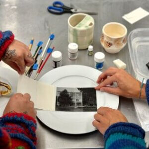 Hands apply a photographic decal to a ceramic plate with tools nearby